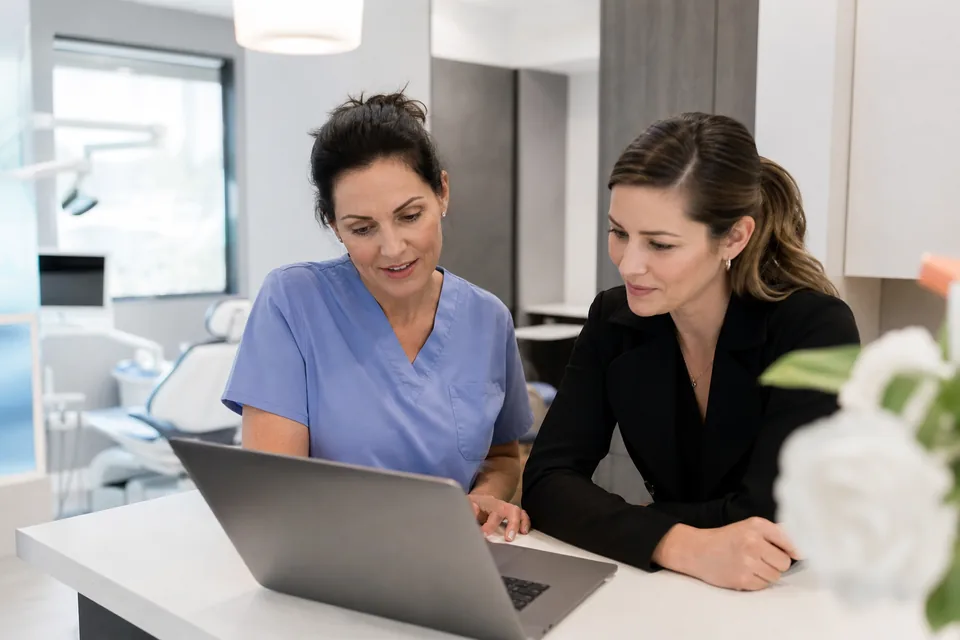 Dentist and office manager reviewing rival dental practices' ads on a laptop at the front desk